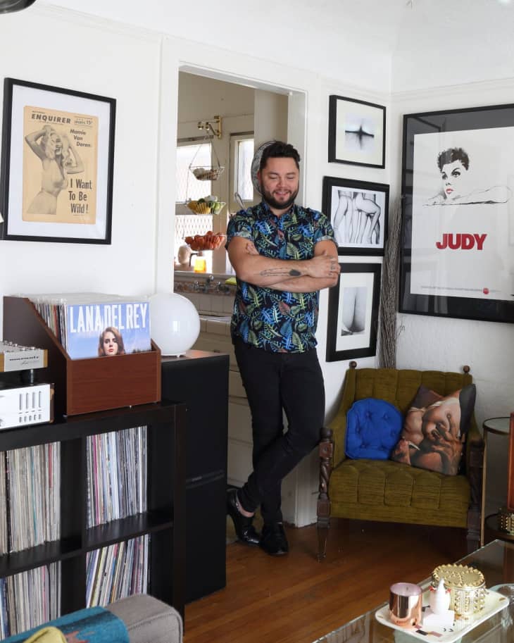 Man in a patterned shirt stands in a living room with framed art, vinyl records, and a green armchair.