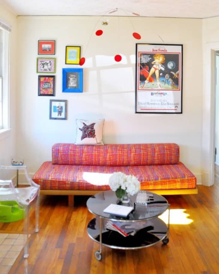 Colorful living room with a red patterned sofa, framed art, a round coffee table, and a clear chair on wooden flooring.