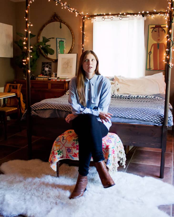 Woman sitting on a bench in a cozy bedroom with a canopy bed, string lights, and eclectic decor.