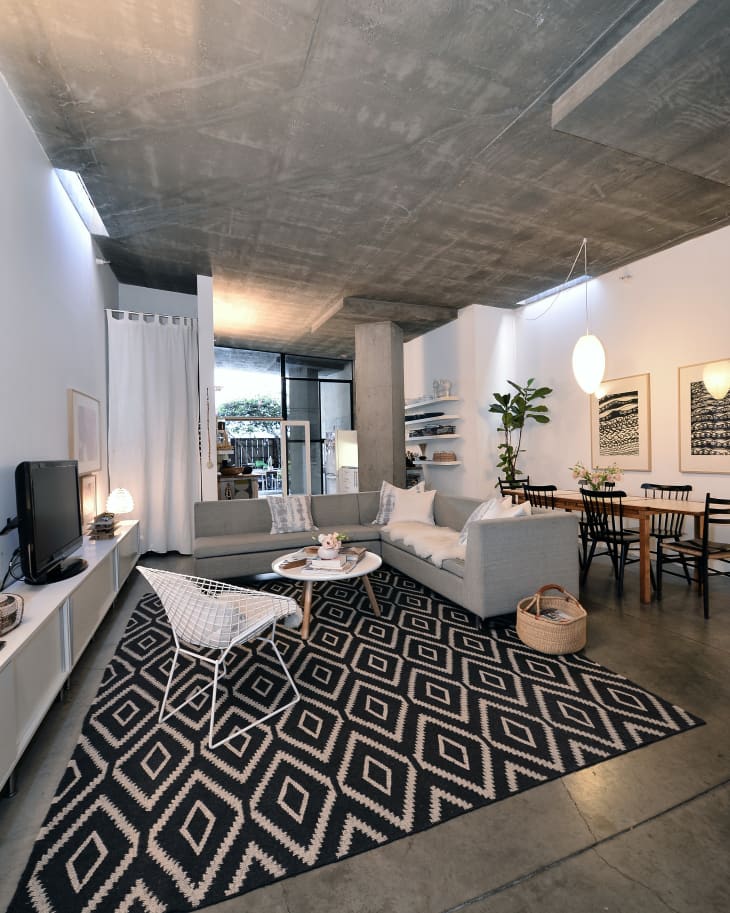 Modern living room with gray sectional sofa, geometric rug, white chair, and dining area with wooden table and black chairs.