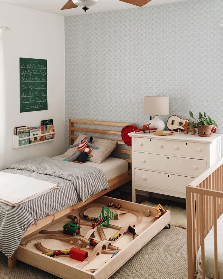 Child's bedroom with wooden bed, pull-out train set, white dresser, and crib. Decor includes a plant and a small guitar.