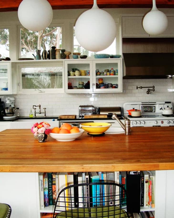 Modern kitchen with wooden island, white cabinets, hanging globe lights, and a bowl of oranges.