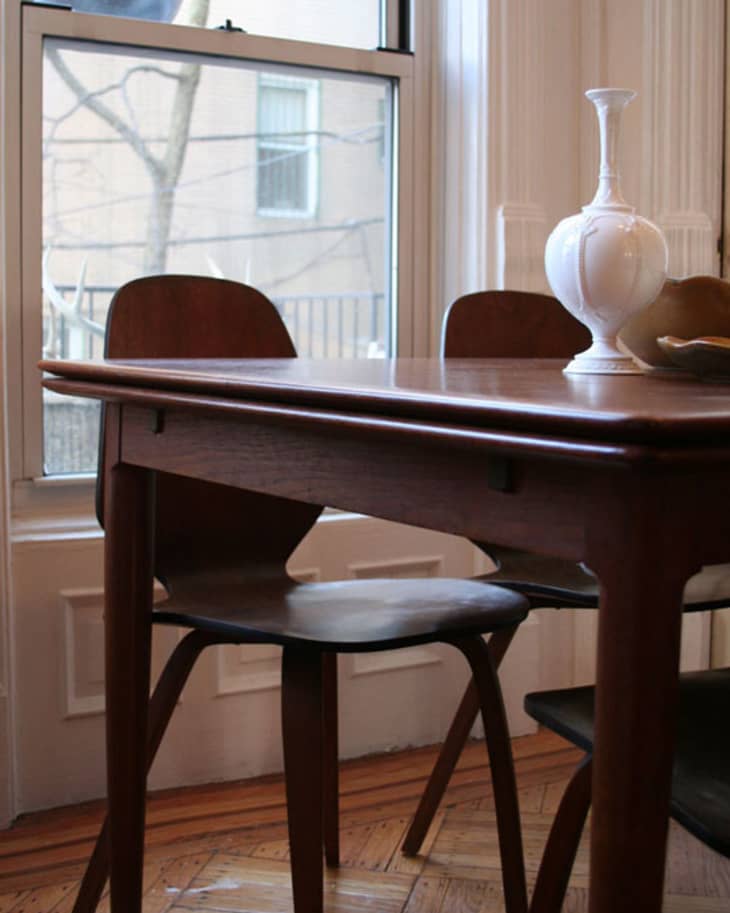 Mid-century modern dining table with two wooden chairs and a white vase by a large window.