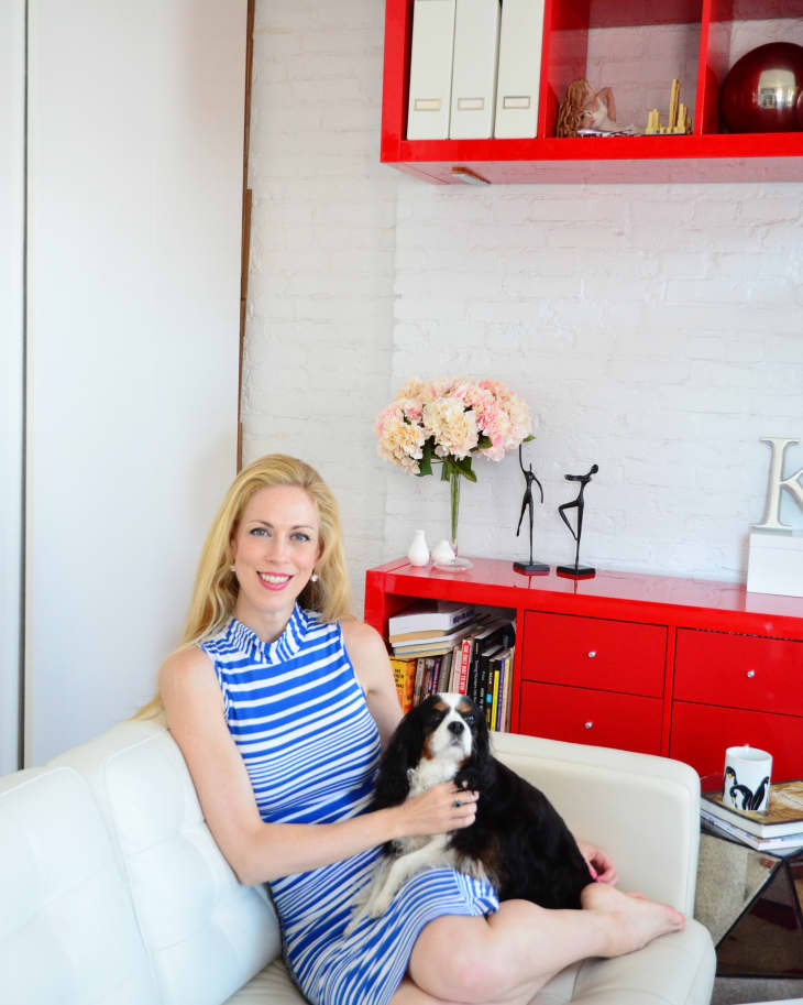 Woman in a blue striped dress sitting on a white sofa with a dog, red shelves and decor in the background.