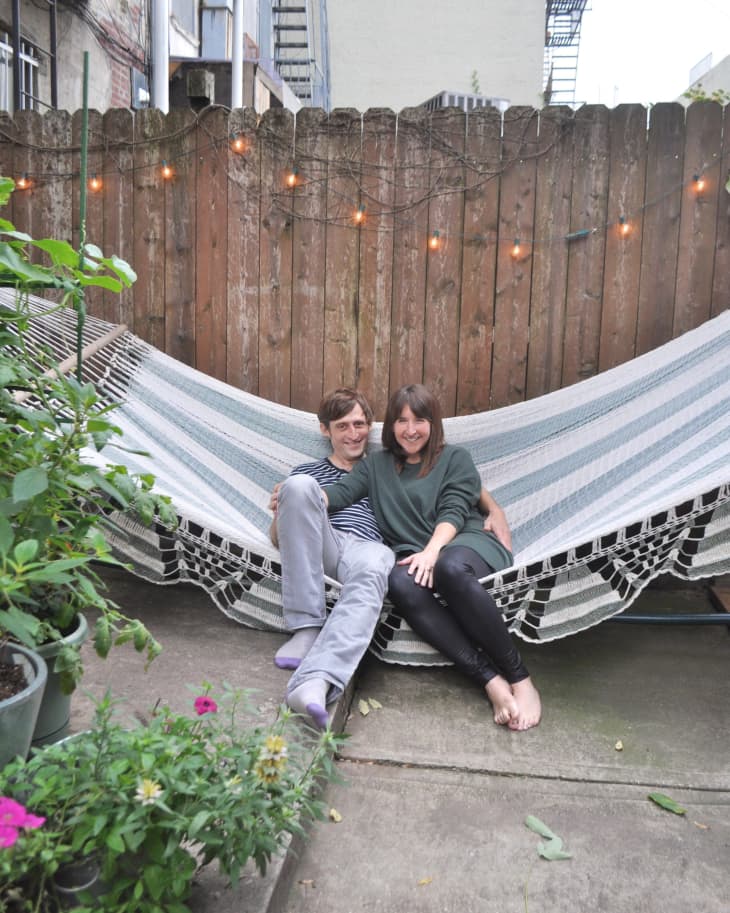 Couple sitting on a striped hammock in a garden with potted plants and string lights on a wooden fence.