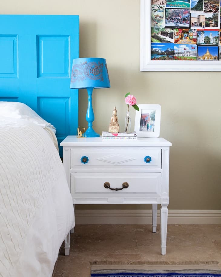 White nightstand with blue lamp, pink flower, and framed photos beside a bed with a blue headboard.