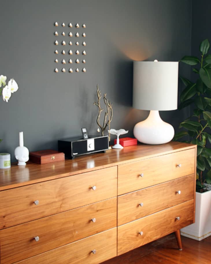 Wooden dresser with white lamp, orchid, and decor on top, against a dark wall with a grid of small metallic knobs.