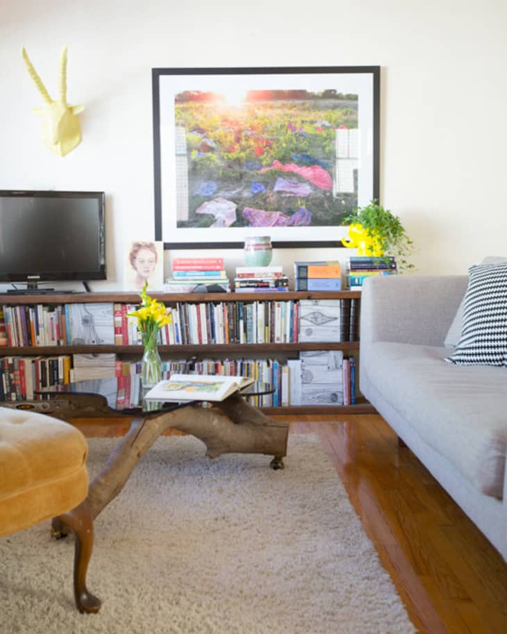 Living room with a gray sofa, wooden coffee table, bookshelf, TV, and framed landscape art. Yellow flowers on the table.