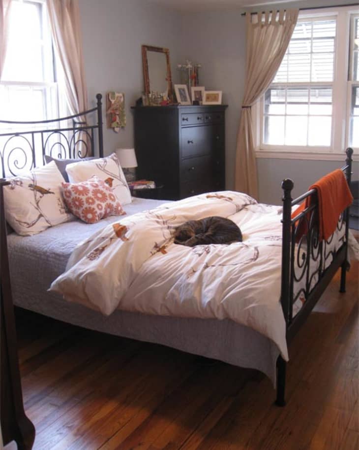 Cozy bedroom with wrought iron bed, floral bedding, and a cat sleeping. Black dresser and framed photos on top.