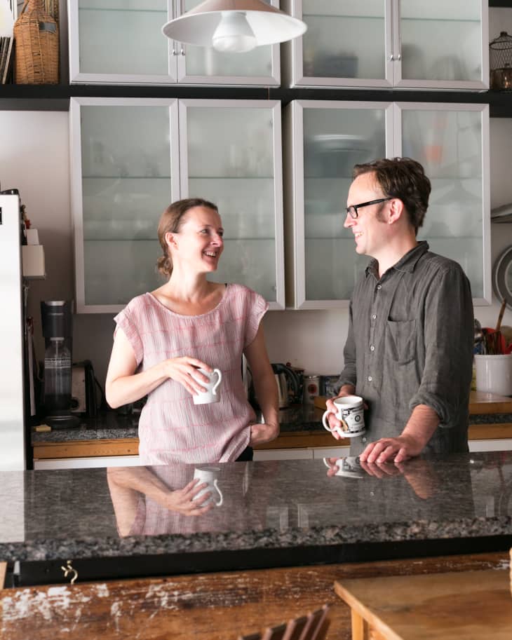 Two people smiling and holding mugs in a modern kitchen with glass cabinets and a pendant light.