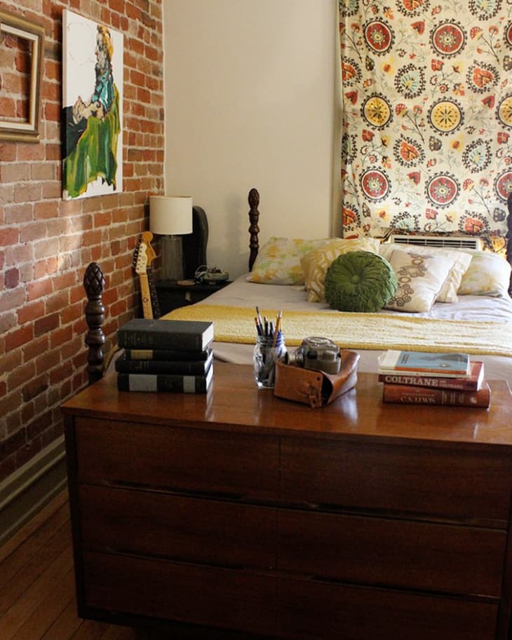 Bedroom with exposed brick wall, wooden dresser, books, and a bed with patterned pillows and a colorful tapestry.
