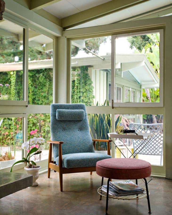 Mid-century modern sunroom with a blue armchair, pink ottoman, and potted plants by large windows.