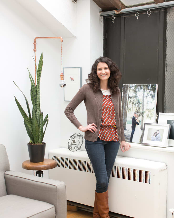 Woman in a living room with a snake plant, gray armchair, and framed photos on a radiator.