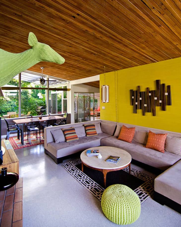 Mid-century modern living room with gray sectional, yellow accent wall, geometric art, and green pouf. Dining area in background.