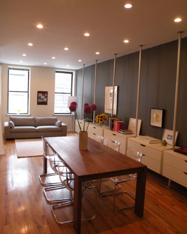 Modern living room with a wooden dining table, clear chairs, gray sofa, and wall-mounted shelves with books and decor.