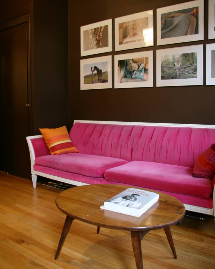 Pink velvet sofa with orange cushions, wooden coffee table, and framed photos on a dark wall.
