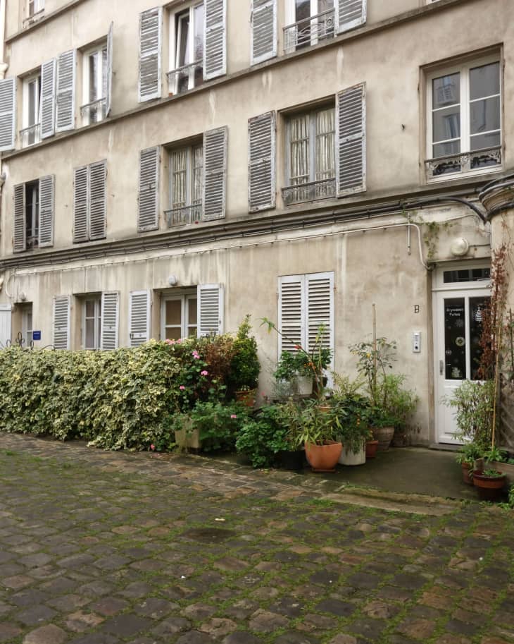 Facade of an old building with shuttered windows, potted plants, and cobblestone courtyard.