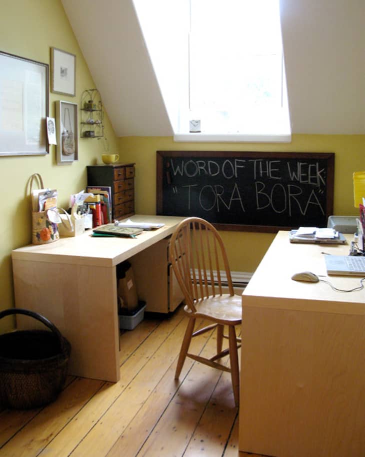 Home office with wooden desks, chair, chalkboard with "Word of the Week: Tora Bora," and framed art on yellow walls.