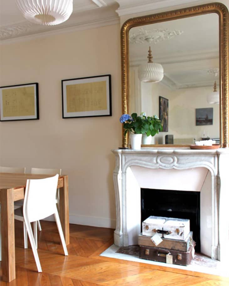 Dining room with wooden table, white chairs, ornate mirror above fireplace, and potted plant on mantel.