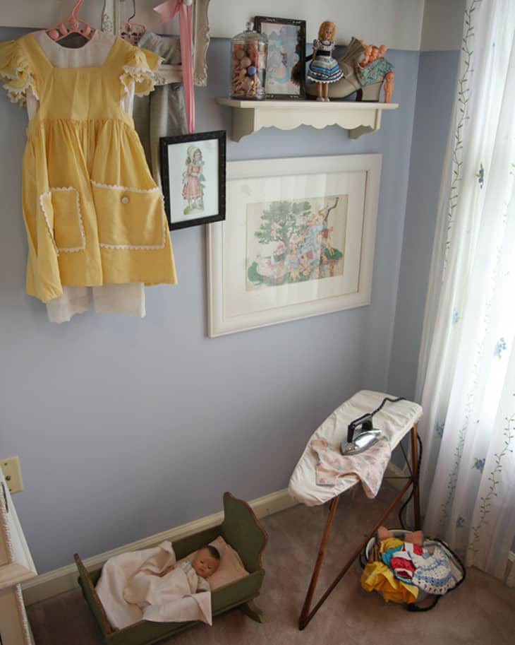 Vintage nursery corner with yellow dress, framed art, dolls, ironing board, and doll cradle on carpet.