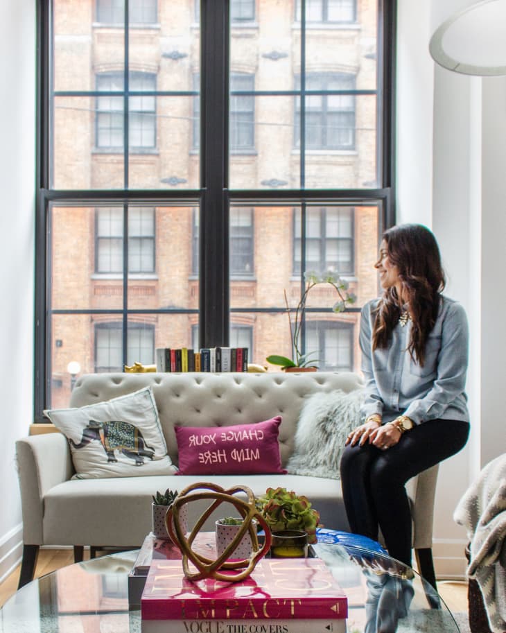 Woman sitting on a sofa with decorative pillows, books, and plants on a coffee table in a bright living room.