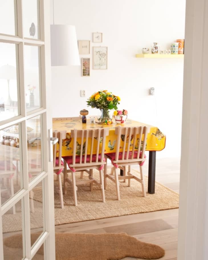 Dining room with a yellow tablecloth, wooden chairs, and a vase of flowers, viewed through open French doors.