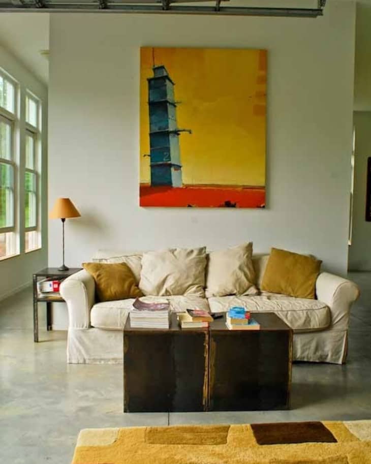 Beige sofa with brown cushions, abstract painting, side table with lamp, and books on a wooden coffee table.