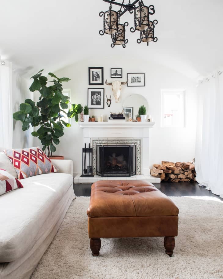 Living room with a white sofa, brown ottoman, fireplace, framed photos, and a large plant.