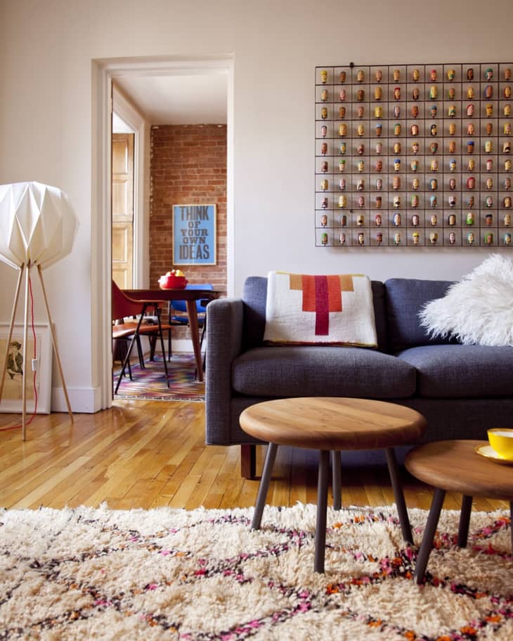 Living room with gray sofa, colorful throw, round wooden tables, and wall art. View into dining area with brick wall.