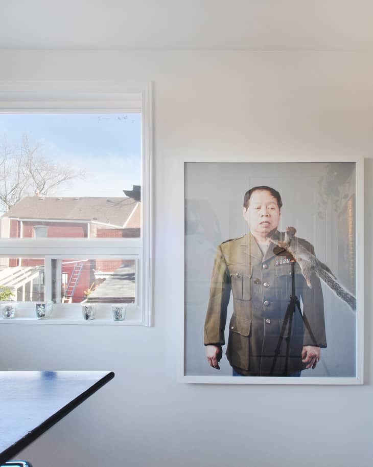 Framed portrait of a man in a military uniform with a bird on his shoulder, next to a window with a view of rooftops.