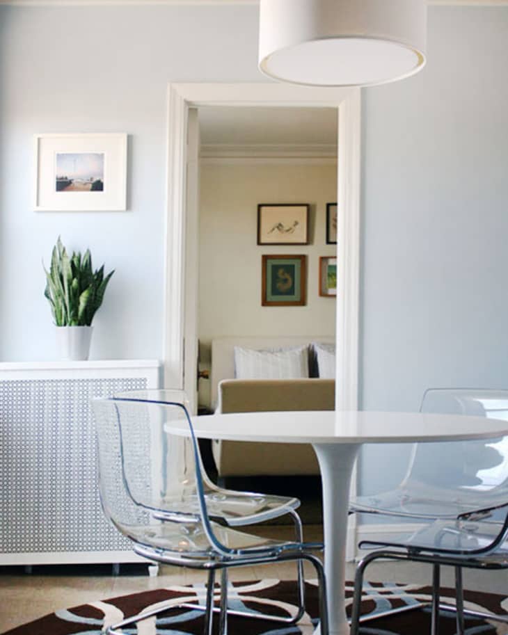 Modern dining area with a white round table, clear chairs, and a geometric rug, leading to a cozy room with framed art.