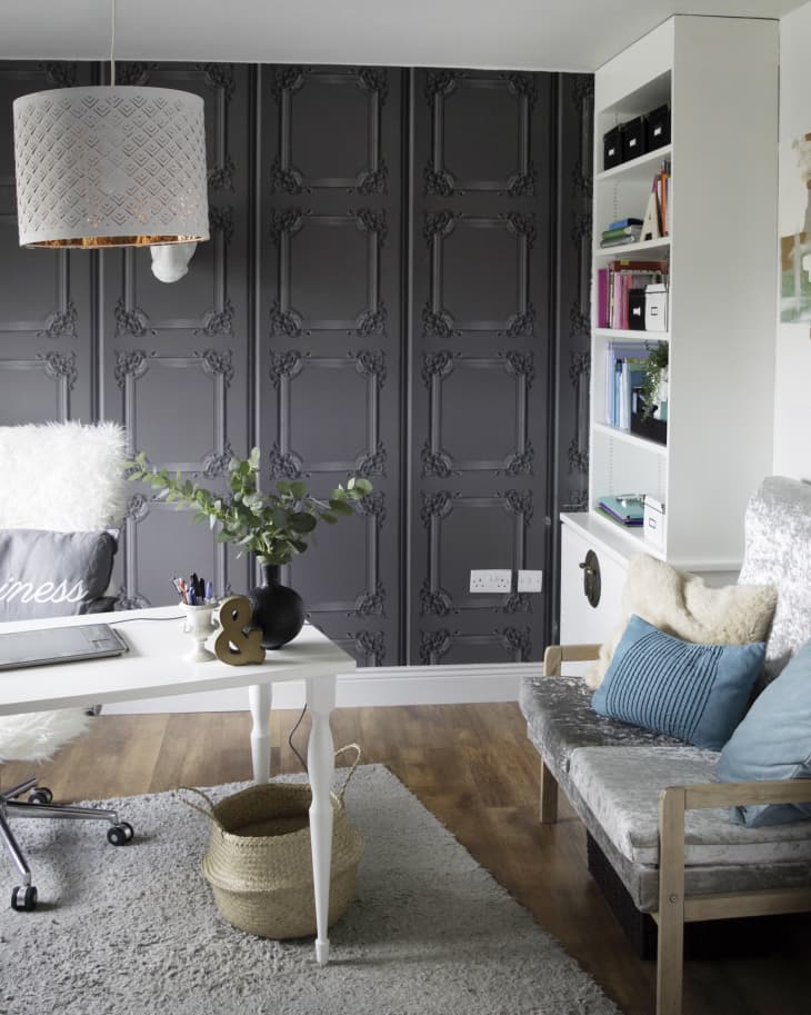 Home office with dark paneled wall, white desk, wicker basket, and cozy chair with blue cushions.