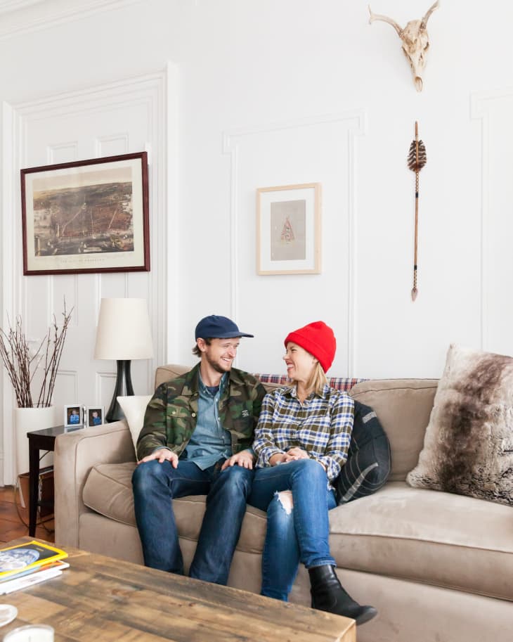 Couple sitting on a beige sofa in a living room with wall art, a skull decor, and a rustic wooden coffee table.