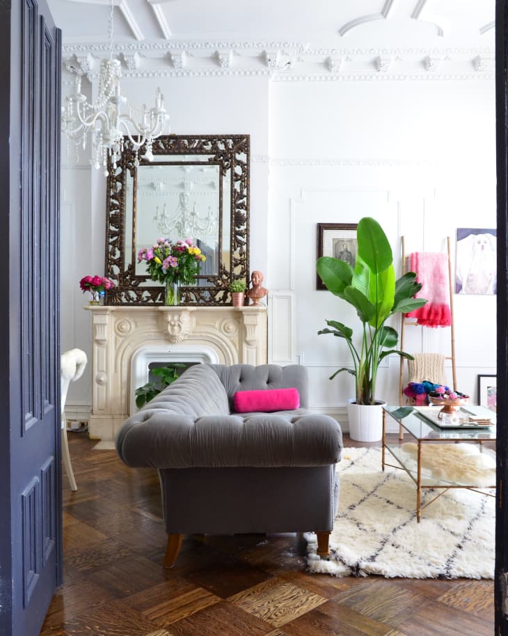 Elegant living room with gray sofa, ornate mirror, chandelier, and vibrant plants on a patterned rug.