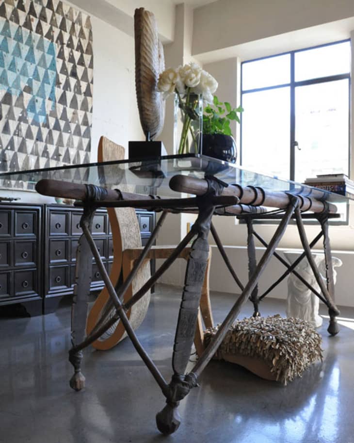 Glass-top table with metal legs, wooden chair, decorative vase with white flowers, and geometric wall art in a sunlit room.