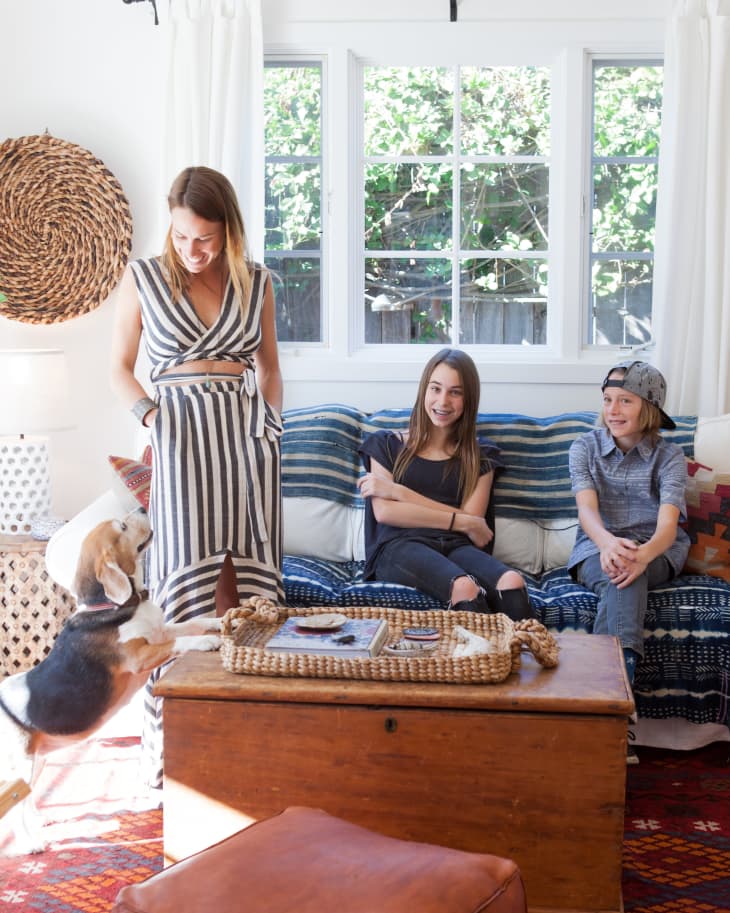 Woman in striped dress with two kids on a sofa, beagle jumping on wooden chest, woven decor on wall.