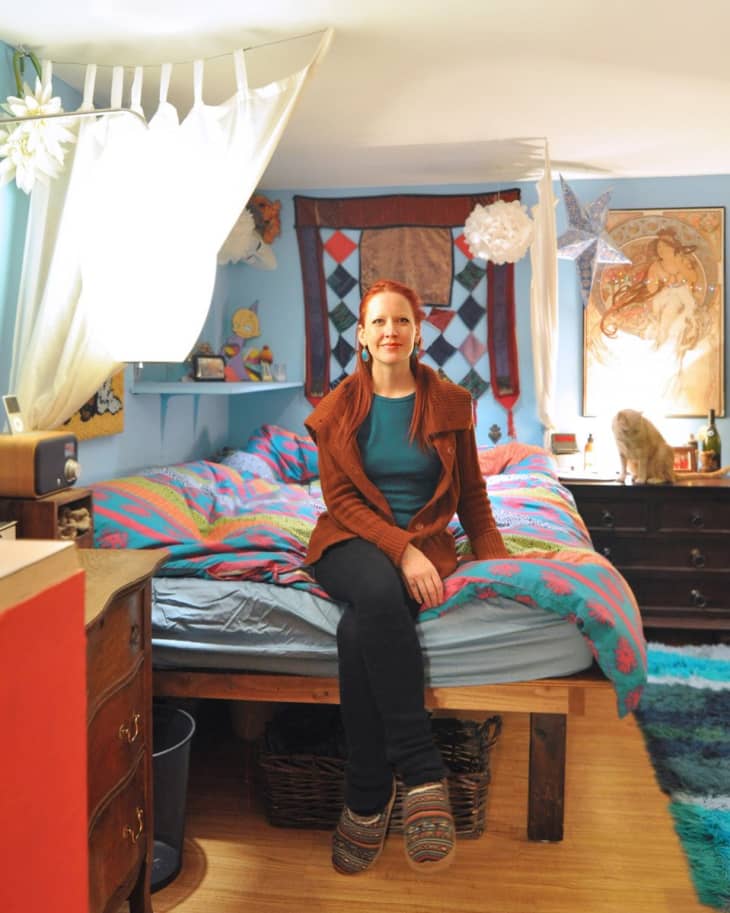 Woman sitting on a colorful bed in a cozy room with blue walls, wooden floor, and eclectic decor.