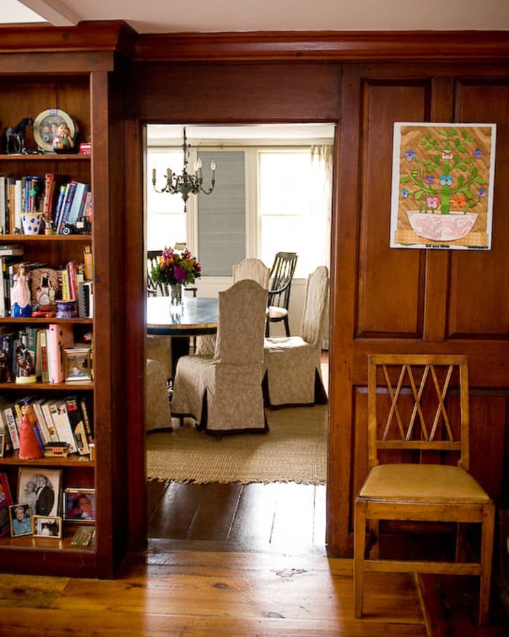 Wood-paneled room with a bookshelf, leading to a dining area with slipcovered chairs, a chandelier, and a floral centerpiece.