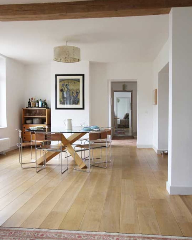 Dining room with glass table, wooden floor, modern chairs, and a sideboard with bottles. Framed art and pendant light above.