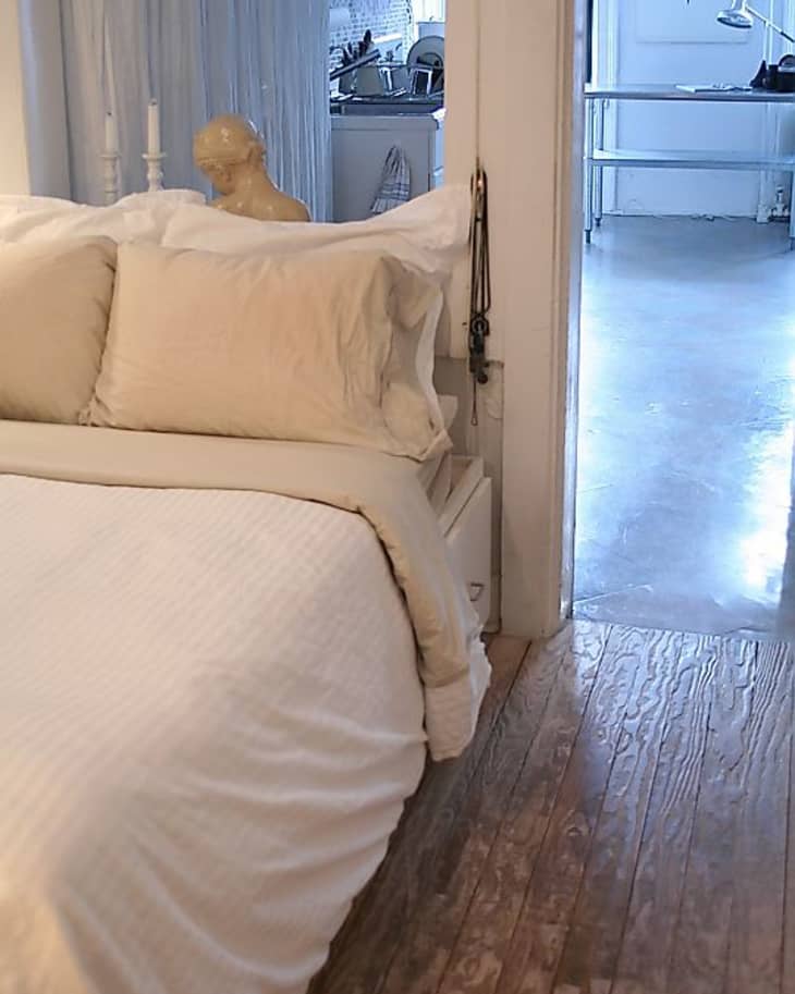 Cozy bedroom with white bedding, wooden floor, and a view into a kitchen and workspace with a desk and lamp.
