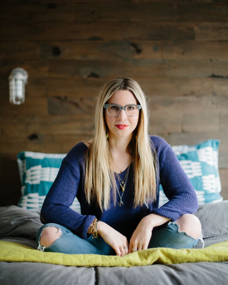 Woman with long blonde hair and glasses sitting on a bed with blue and green pillows, wearing a blue sweater and ripped jeans.