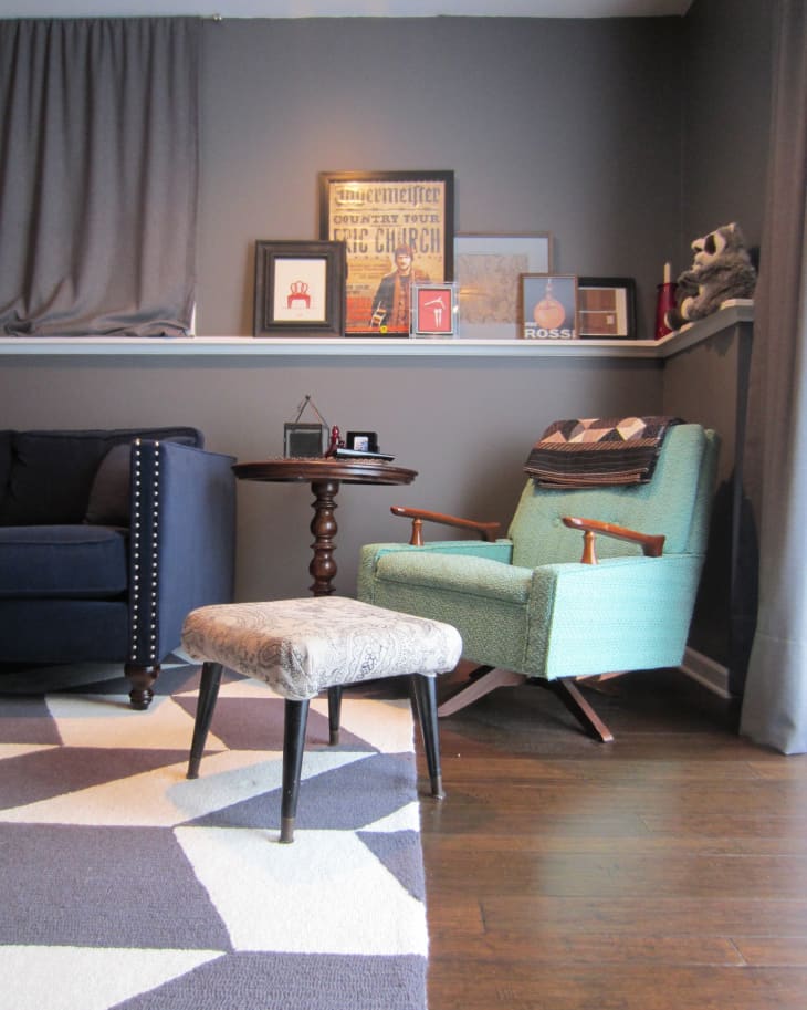 Living room with a blue sofa, teal armchair, patterned ottoman, and framed art on a shelf against a gray wall.