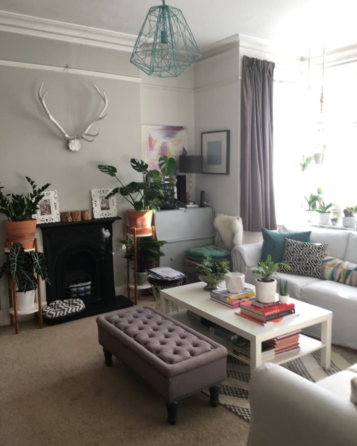 Living room with white sofa, tufted ottoman, plants, books on coffee table, and antler wall decor above fireplace.