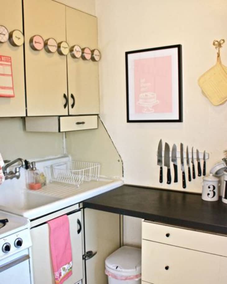 Small kitchen with cream cabinets, wall-mounted knives, dish rack, and pink towel.