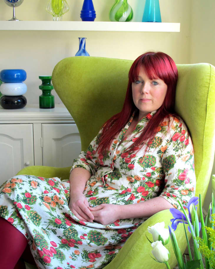 Woman with red hair in floral dress sitting on a green armchair, surrounded by colorful glass vases and flowers.