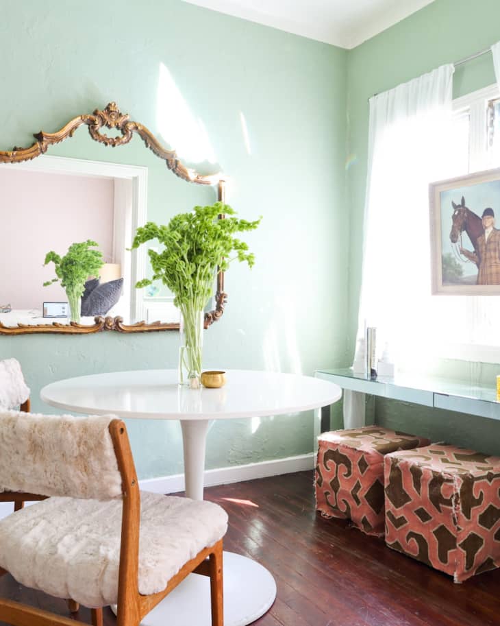 Dining area with a white round table, green walls, ornate mirror, and a vase of greenery.