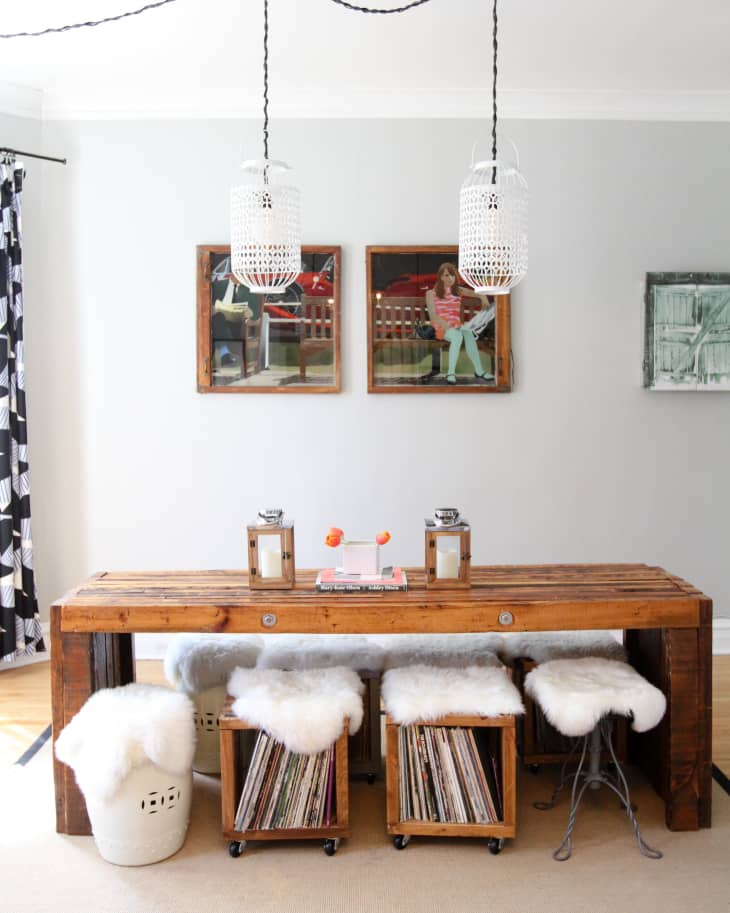 Wooden dining table with white fur stools, hanging lanterns, and framed art on a gray wall. Vinyl records stored below.