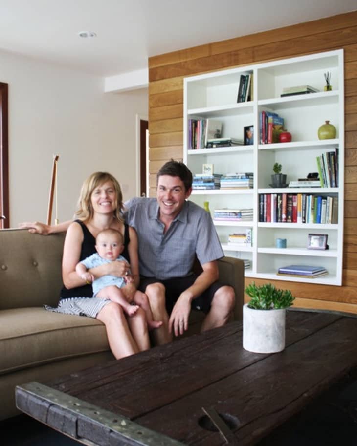 Family sitting on a beige sofa in a living room with a wooden wall and white bookshelf filled with books and decor.