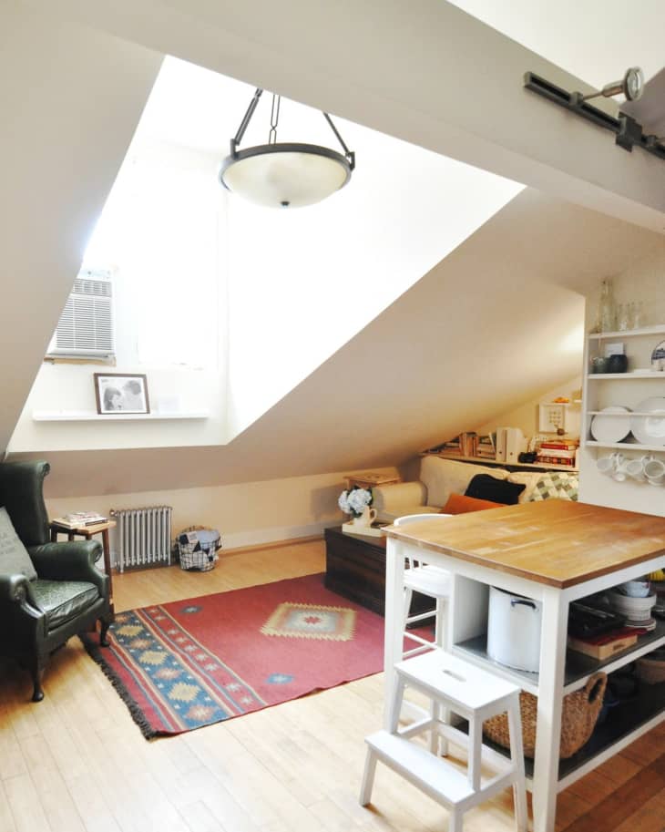 Attic room with sloped ceiling, red patterned rug, green armchair, wooden table, and shelves with dishes and books.