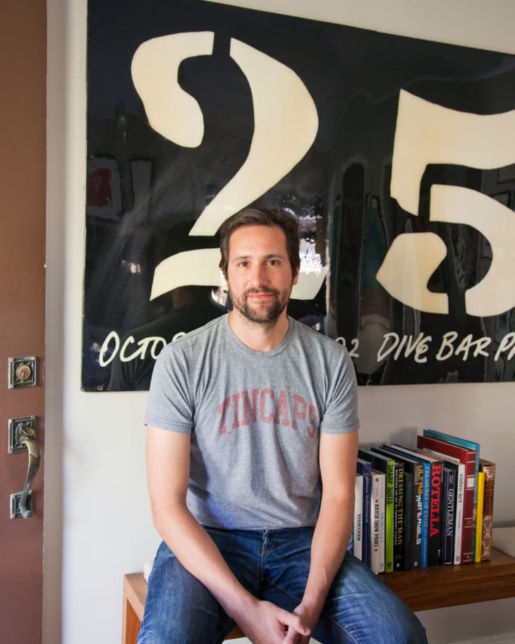 Man in a gray T-shirt sitting on a bench, with books and a large "25" sign in the background.